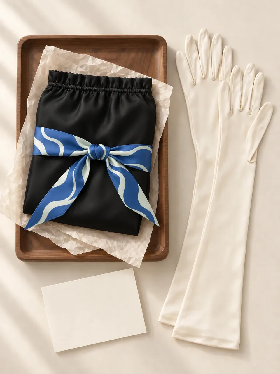 A black silk sleep bonnet tied with a blue skinny silk scarf beside ivory silk gloves on a walnut tray