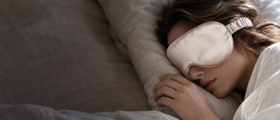 A woman asleep on linen pillows wearing a champagne silk sleep mask